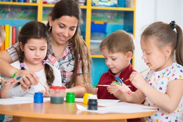 A childcare provider teaches three young children to paint.